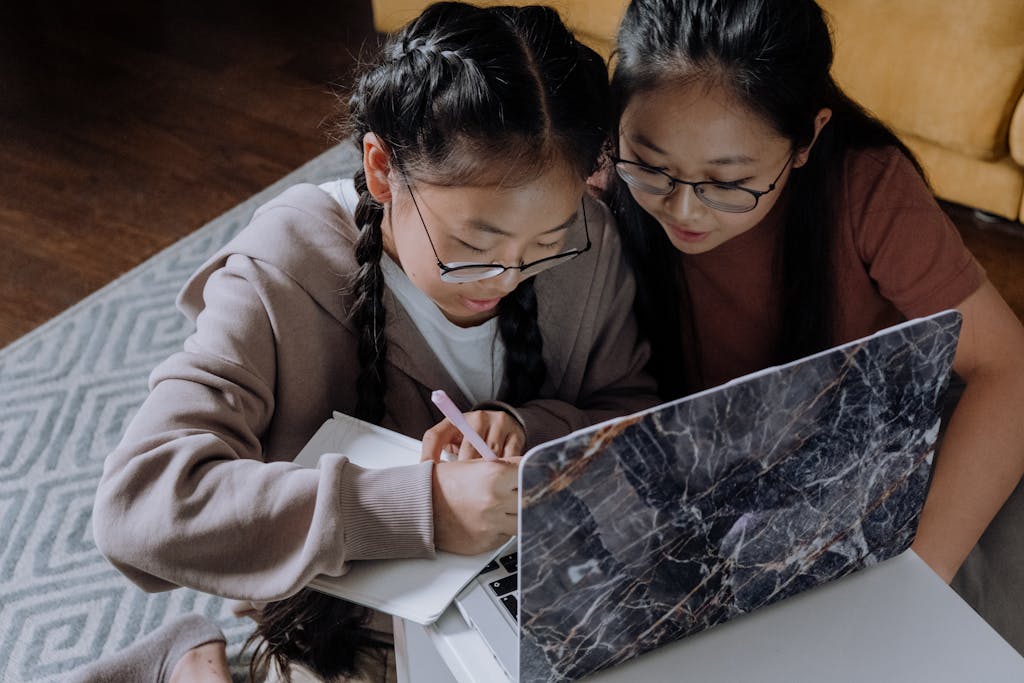 Two sisters studying together at home, using a laptop and writing in a notebook, fostering learning and family bonding.